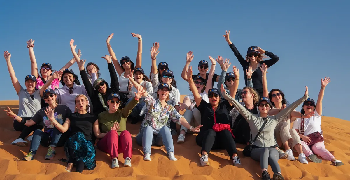 Large group of people sitting together on a sand dune, raising their arms and posing cheerfully under a clear blue sky.
