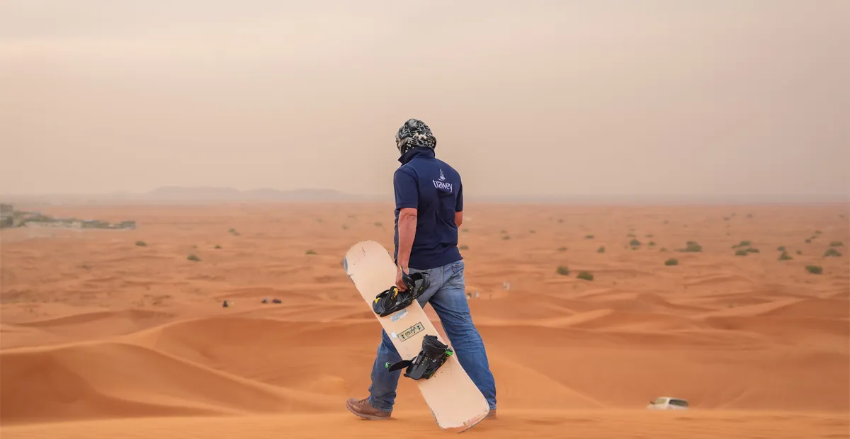 A person wearing a Trawey shirt stands on a sand dune, holding a sandboard, overlooking a vast desert landscape with scattered shrubs and distant vehicles under a hazy sky.