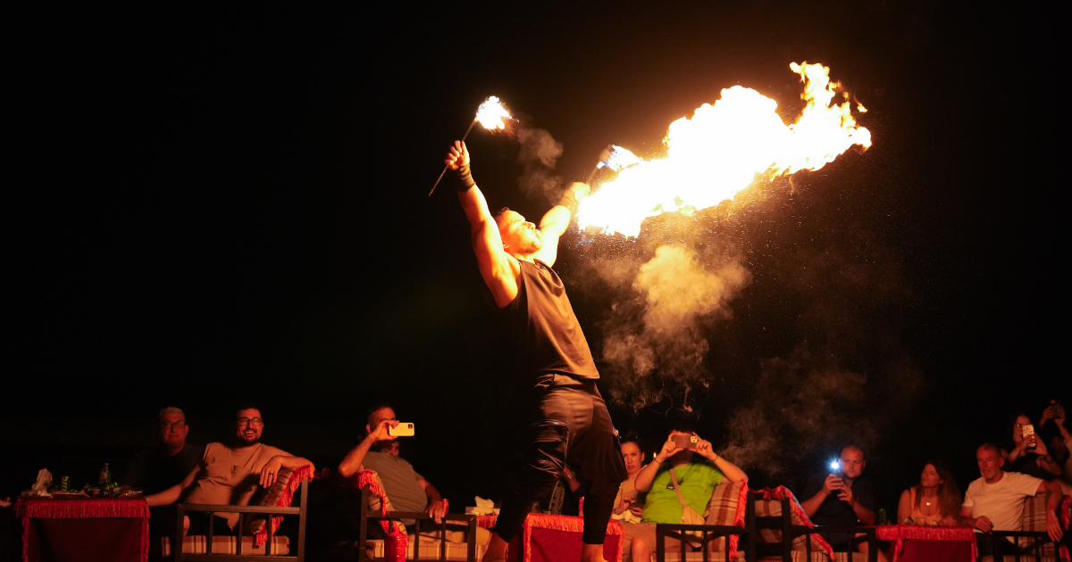 A fire performer blows a large flame on stage at a desert overnight camp, entertaining an audience seated outdoors at night.