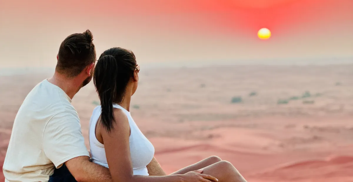 Couple sits together on a sand dune, watching the sunset over a vast desert landscape.