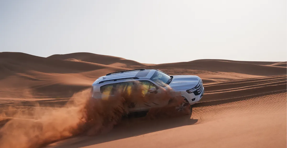 White SUV drives through sand dunes, kicking up orange sand.