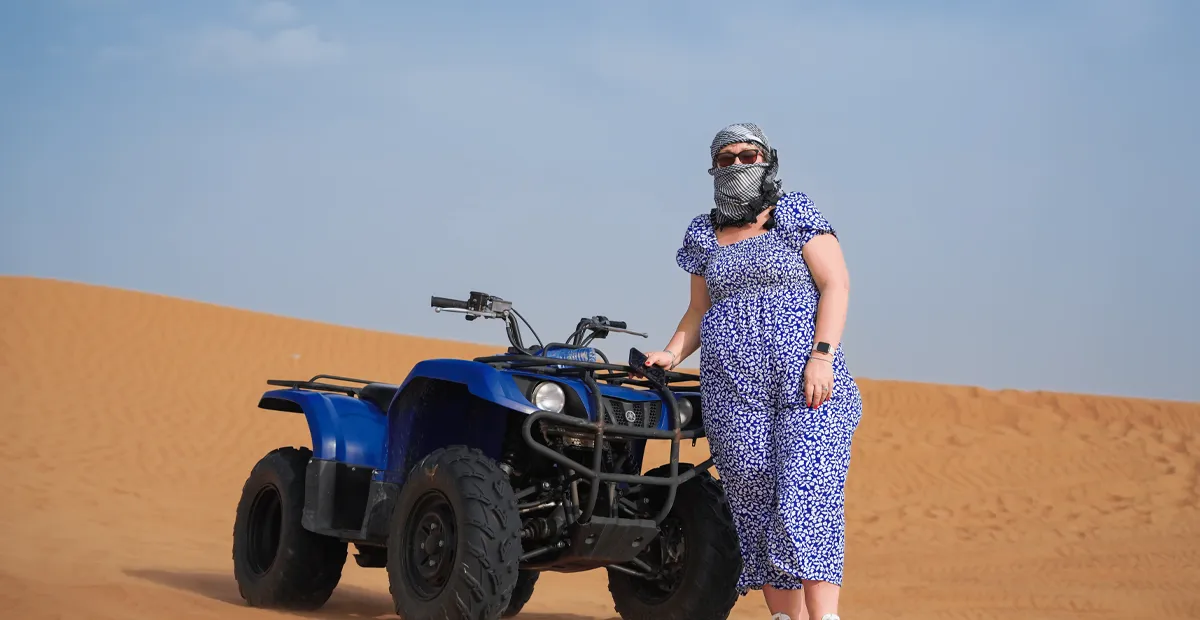 Person in a blue outfit stands beside a blue quad bike on desert sand dunes under a clear sky.