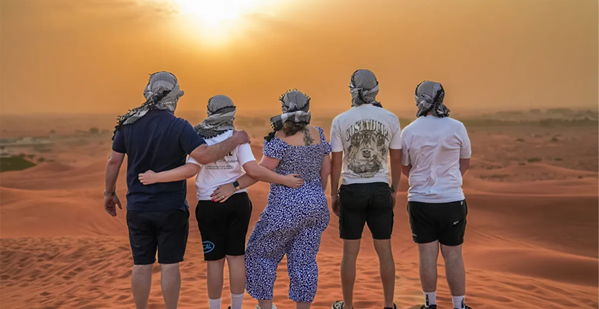 Five people wearing headscarves stand arm in arm on a sand dune, watching the sunset over the desert.