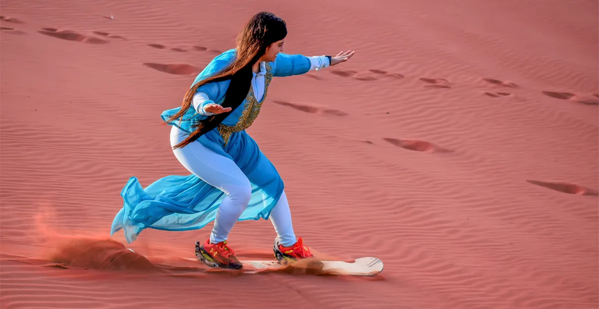 Woman in a blue traditional outfit sandboarding down a red desert dune, leaving a trail in the sand.