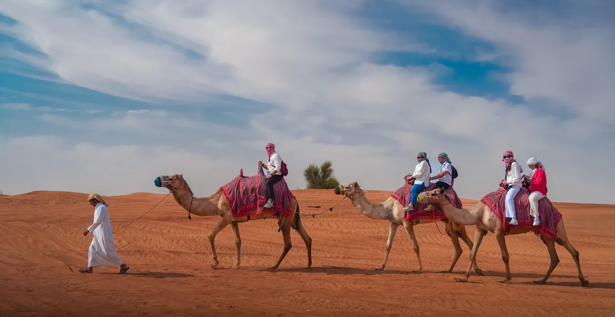 Group of people riding blue quad bikes across orange sand dunes under a clear blue sky.