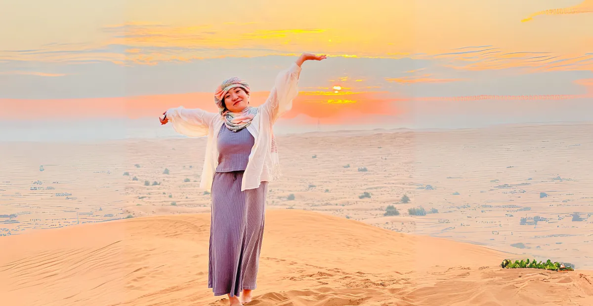 Person in a long dress and scarf stands barefoot on a sand dune at sunset, arms raised, with a vast desert landscape in the background.