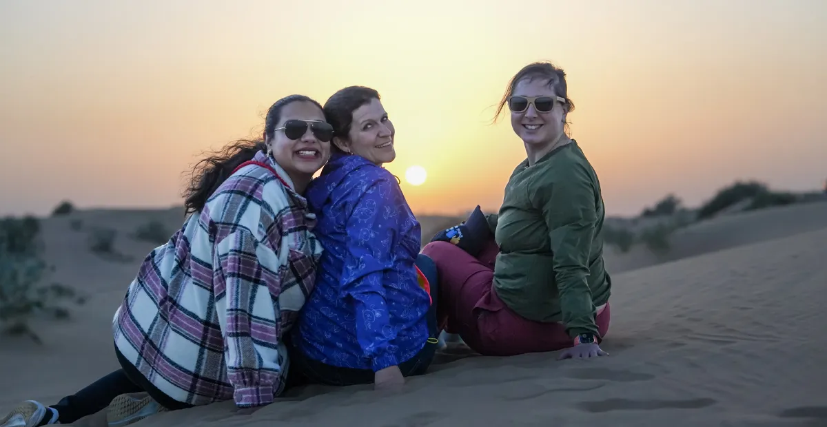 Three people sit together on a sand dune at sunset in the desert, with the sun low on the horizon.