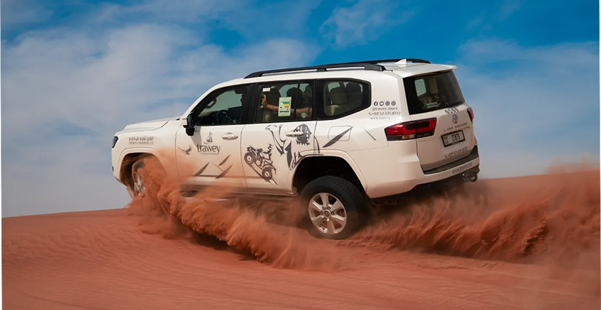 White SUV drives up a red sand dune, kicking up clouds of sand under a blue sky.