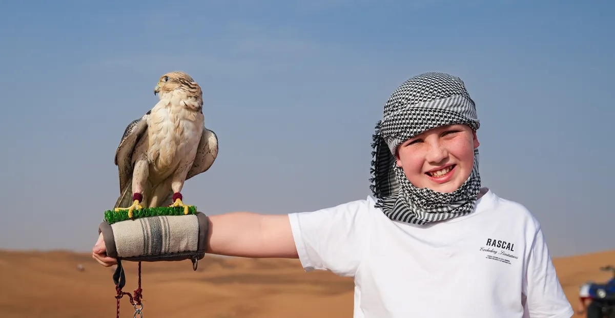 Person wearing a keffiyeh and white t-shirt holds a falcon on a gloved arm in the desert, with sand dunes and blue sky in the background.