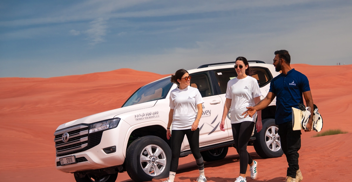 Three people in activewear stand beside a white SUV with "Trawey Tours" branding, parked on red sand dunes under a blue sky, holding sandboards and preparing for a desert safari adventure.