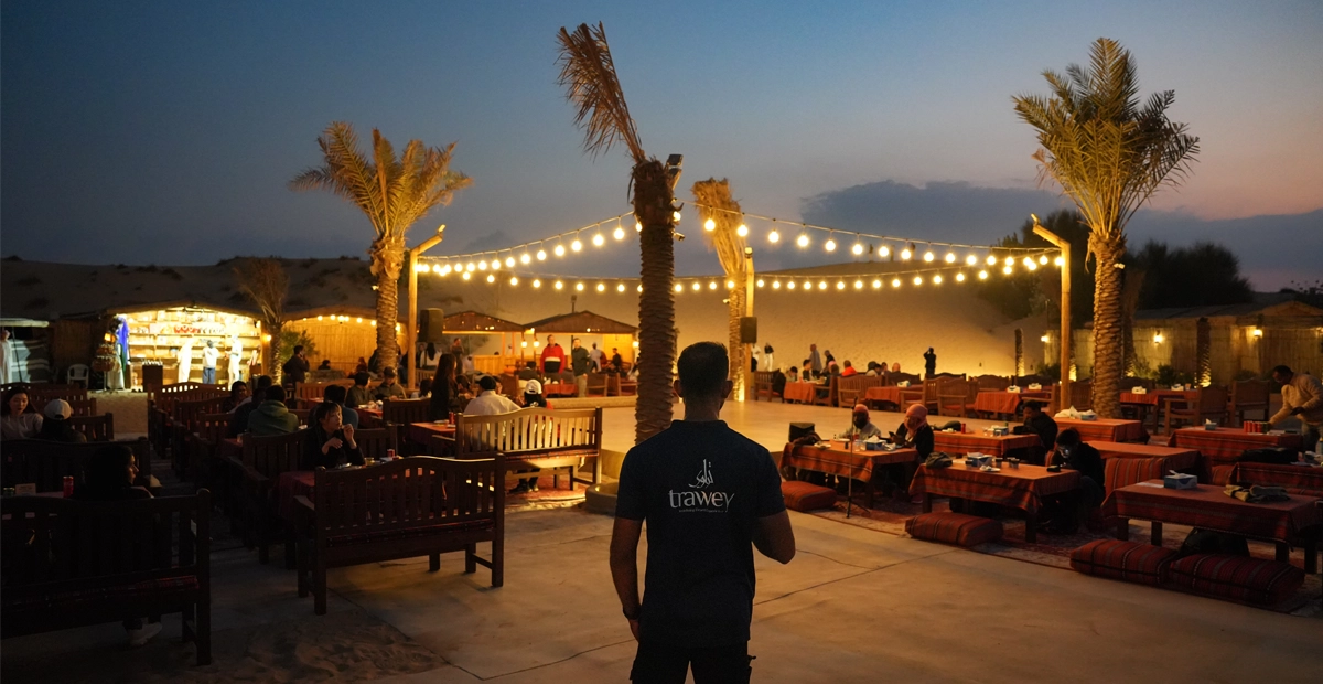 A Dubai desert camp at dusk with guests dining on traditional low tables and benches under string lights and palm trees, surrounded by sand dunes. A staff member in a “trawey” shirt stands in the foreground, capturing the welcoming and festive atmosphere of an evening desert safari.