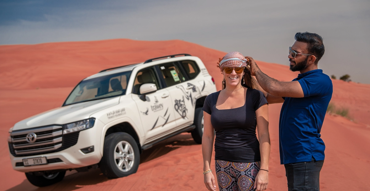 Two people on red desert sand stand in front of a white SUV. One, in a navy shirt and black pants, helps the other, in a black top and patterned leggings, tie a scarf. The setting highlights a desert safari adventure.