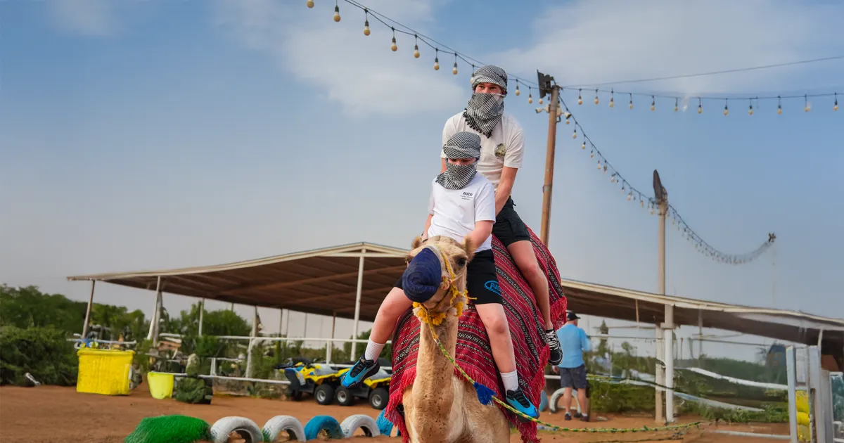 A man and a child, both wearing traditional headscarves, are riding a camel together at a desert safari camp. They are smiling as they ride. The camp's string lights, a shelter, and quad bikes are visible in the background.