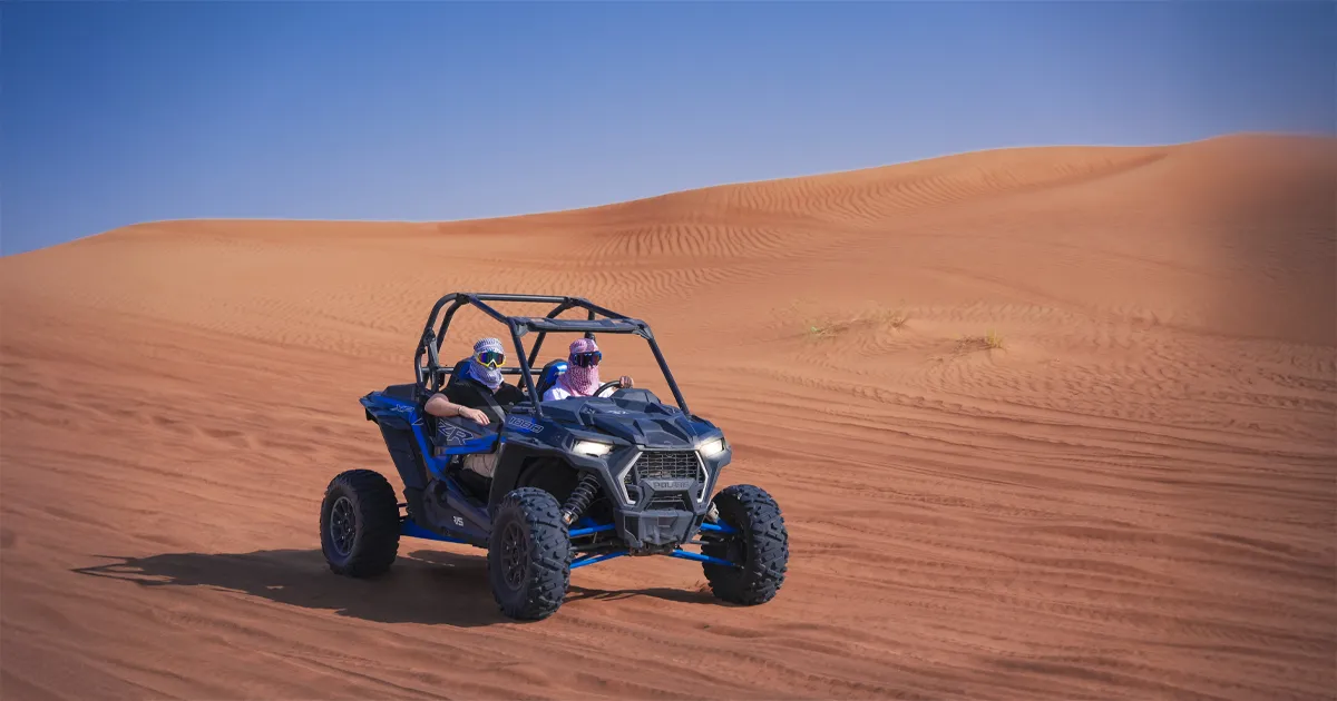Two people in a blue dune buggy are driving on a large red sand dune under a clear blue sky.
