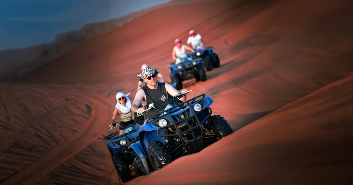 A family is riding blue quad bikes on a steep red sand dune in the desert. The man in the foreground is wearing a black tank top and a bandana, leading the group.