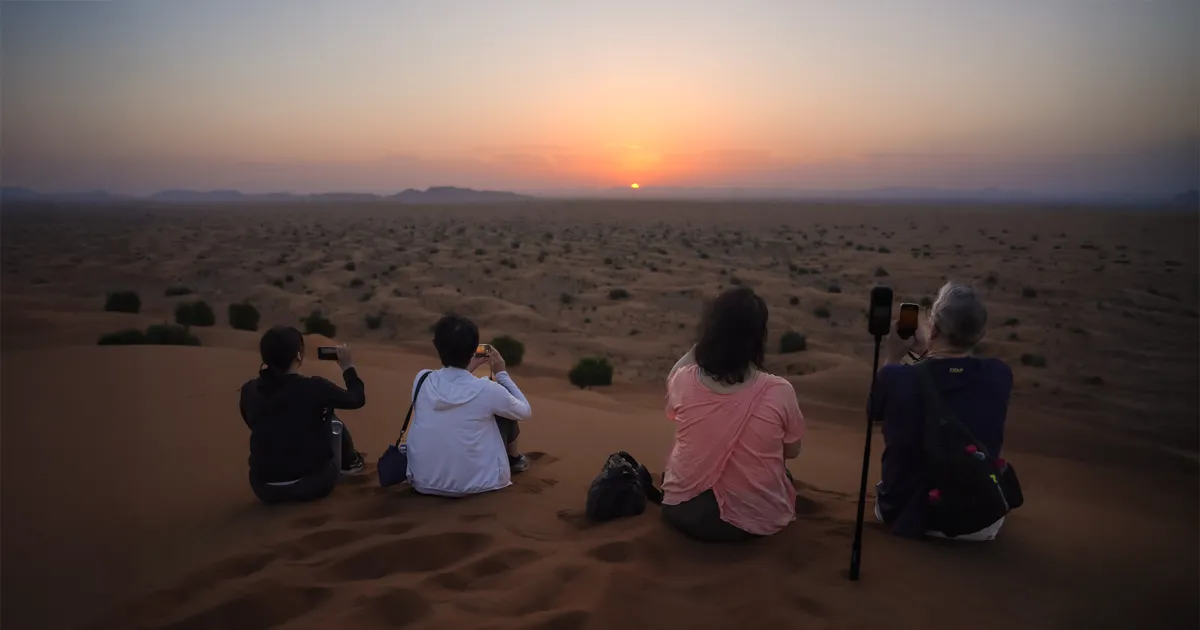 A group of four people sits on a sand dune, taking photos of the sunrise over a vast desert landscape.