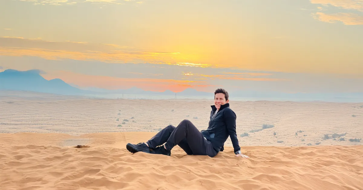 A man sits on a sand dune at sunrise in the desert, wearing a black jacket and black pants. Mountains are visible in the distance under a colorful sky.