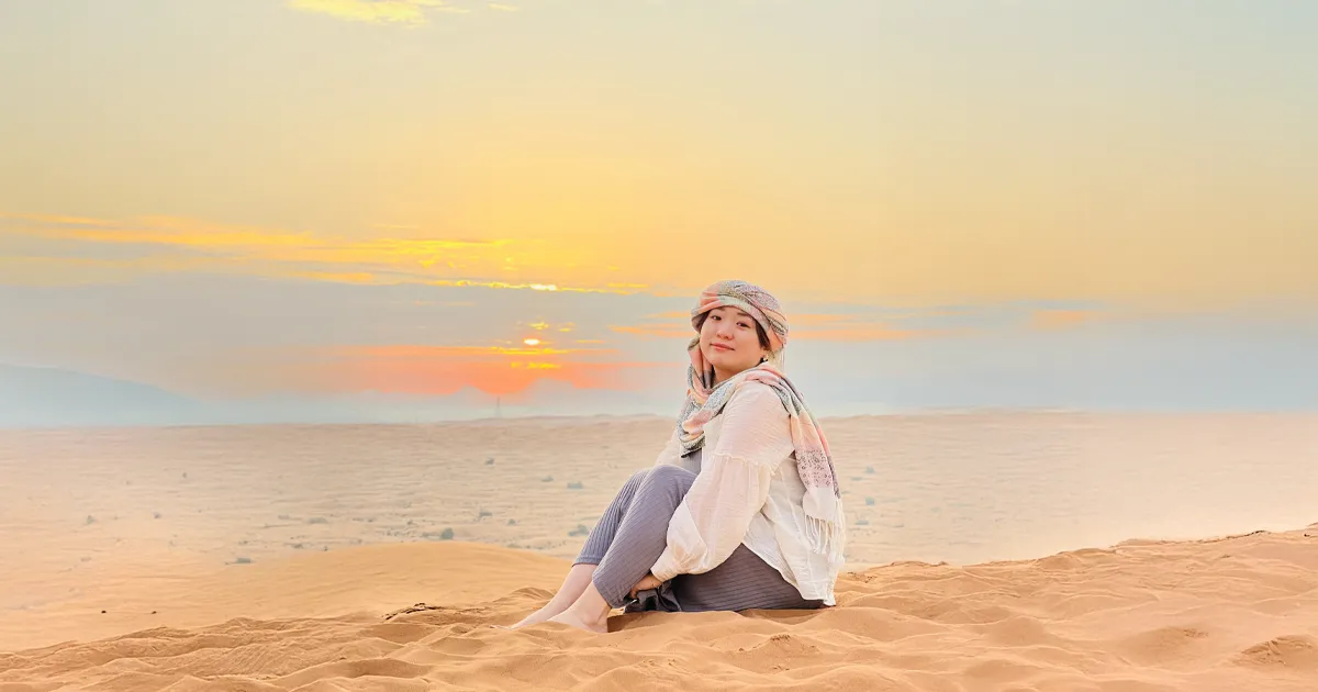 A pregnant woman sits barefoot on a sand dune, smiling and looking at the camera. She wears a white blouse, gray pants, and a headscarf with pink, orange, and green patterns, as the sun sets over the desert landscape behind her.