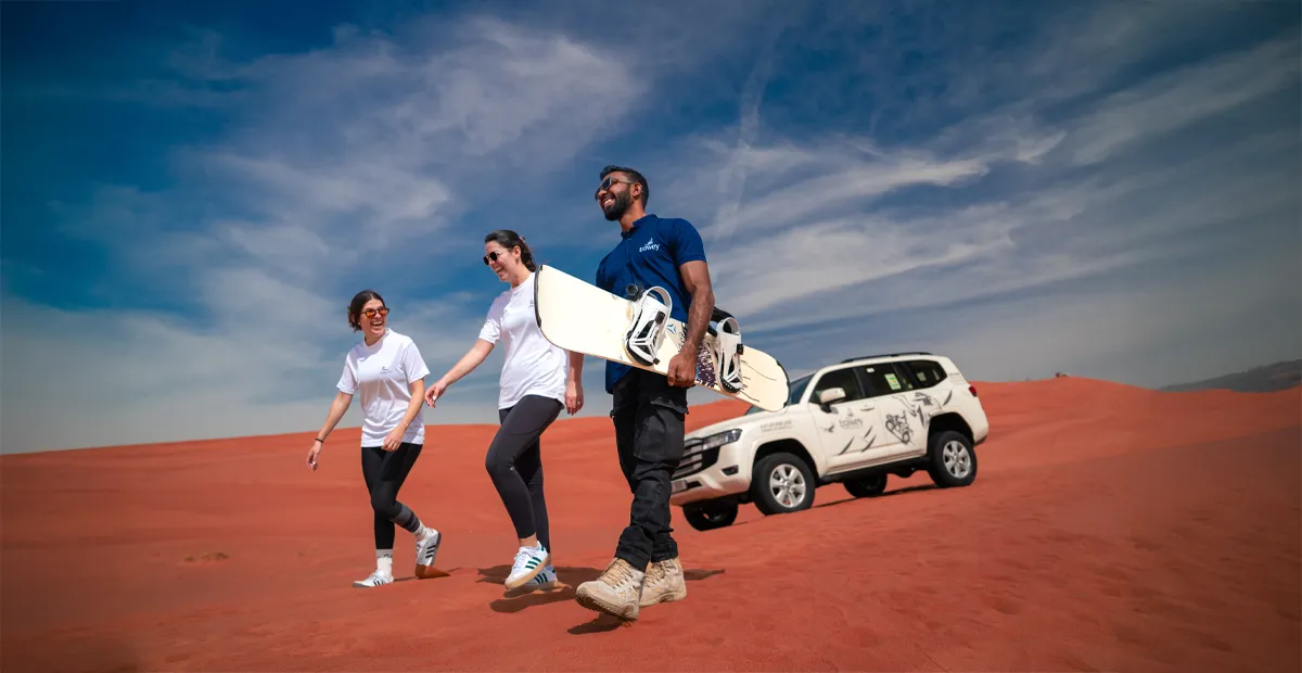 A guide carrying a sandboard walks with two smiling tourists across a red sand dune, with their white 4x4 vehicle parked in the background.