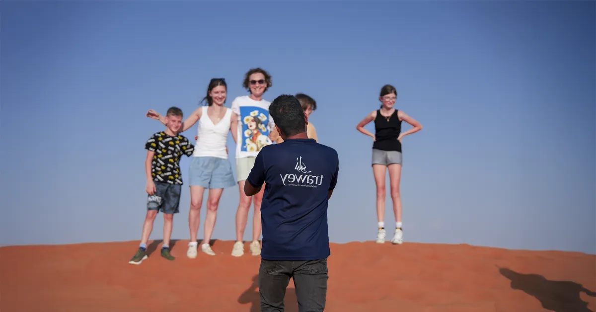 A smiling family of European tourists posing for a photo on a red sand dune, taken by their Trawey guide during a fun desert safari in Dubai.