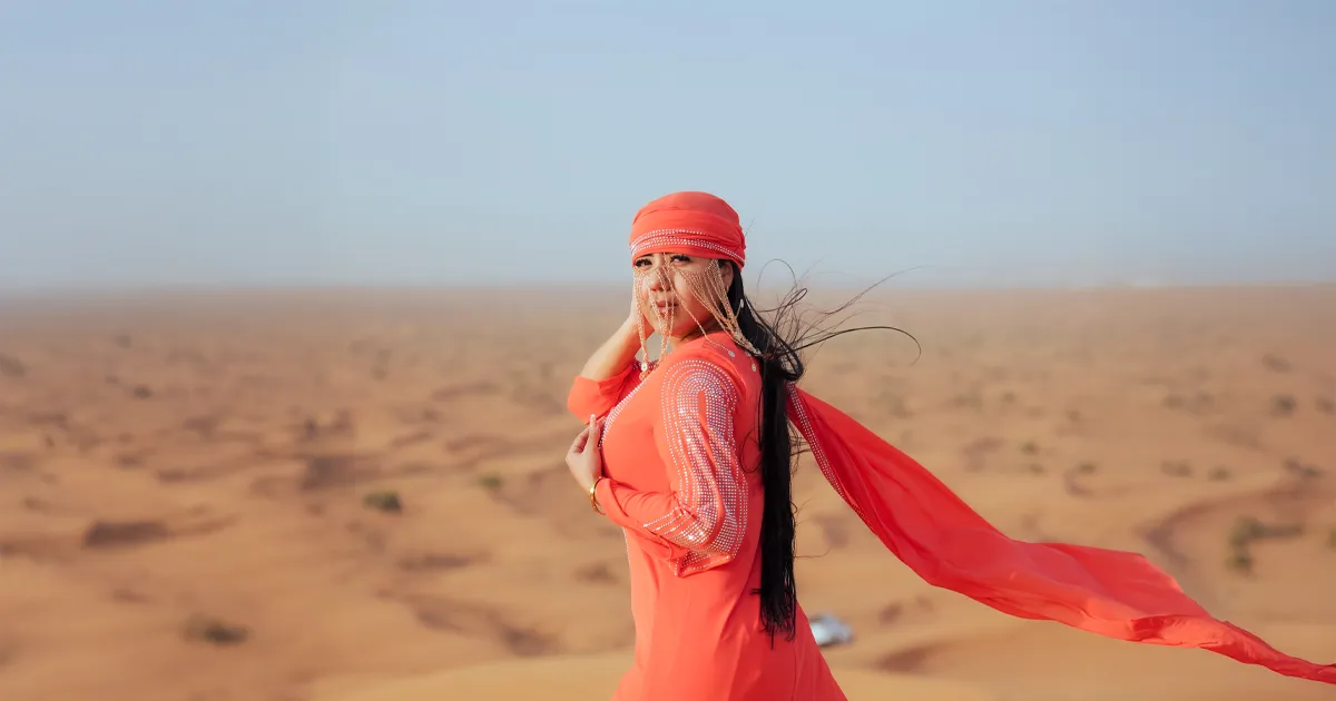 A woman in a vibrant orange traditional dress and headscarf stands on a sand dune, embodying the beauty of Dubai desert sunrise photography.