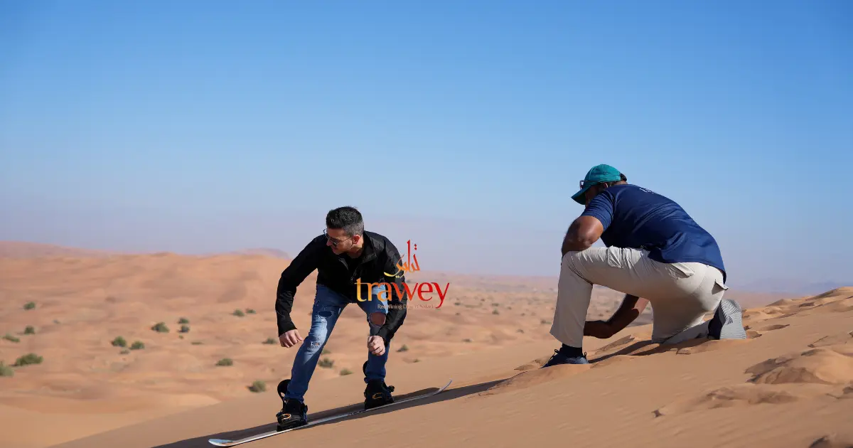 A tourist learns how to go sand boarding in Dubai as an instructor in a blue shirt guides him on a sunny desert dune.