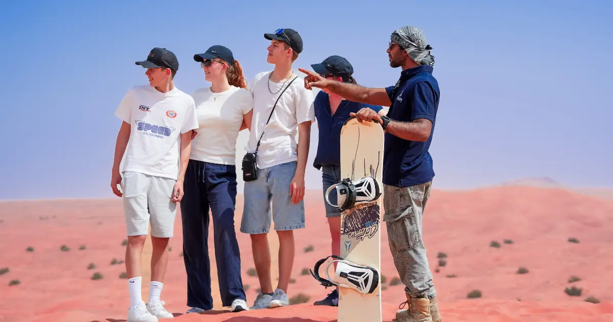 An expert Trawey Tours guide gives a sandboarding safety briefing to a family on a red sand dune in Dubai, holding a board and pointing to the slope.