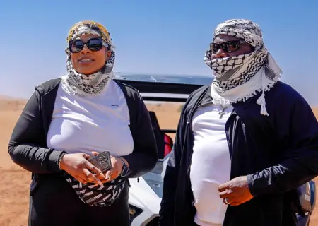 A man and a woman stand side-by-side in a bright, sunny desert landscape. Both are wearing white t-shirts, black zip-up jackets, dark sunglasses, and black-and-white patterned keffiyehs on their heads. The man's keffiyeh is also wrapped around the lower half of his face. They stand in front of a vehicle with sand dunes stretching into the background under a clear blue sky.