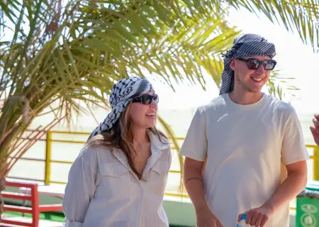 A man and a woman stand side-by-side outdoors, smiling under the bright green fronds of a palm tree. Both are wearing dark sunglasses and black-and-white patterned keffiyehs on their heads.