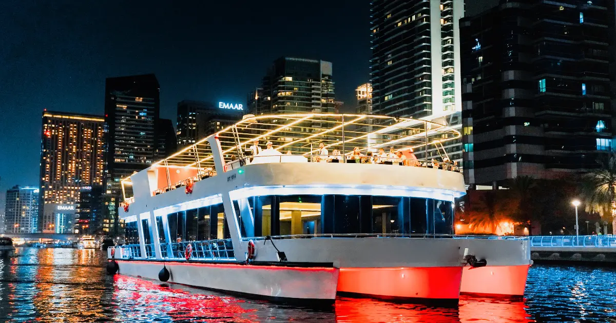 A luxury dinner cruise yacht sailing through Dubai Marina at night, with the illuminated Emaar skyline and city lights reflecting on the water.