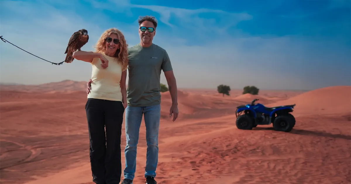 A couple stands in the desert, with a woman holding a falcon on her outstretched arm while a blue quad bike is parked on the sand dunes in the background under a bright blue sky.