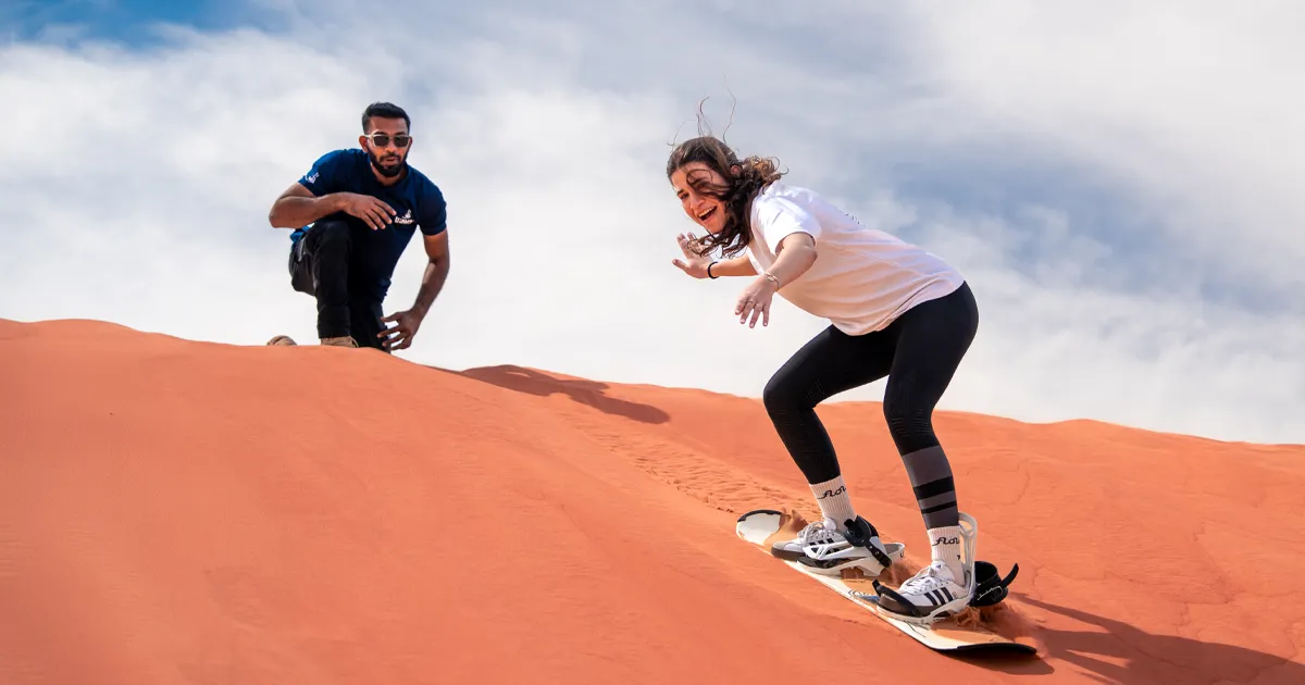 A smiling woman sandboards down a vibrant red sand dune, leaning forward for balance in white sneakers and bindings, while a male instructor in a blue shirt crouches behind her, attentively guiding her technique.