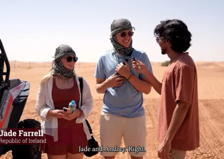 a desert interview shows three people standing in the sand. On the left, a woman with blonde hair and sunglasses wears a red dress, a black and white keffiyeh (headscarf), and a black bag slung over her shoulder. In the center, a tall man with sunglasses also wears a keffiyeh with a light blue t-shirt and beige shorts, holding what appears to be a microphone. On the right, a man with dark, curly hair and glasses, wearing a salmon-colored t-shirt, holds a microphone up to the man in the center.