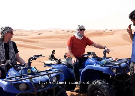 In a bright desert setting with sand dunes in the background, two people sit on blue quad bikes (ATVs). The person on the left wears a keffiyeh (headscarf) and sunglasses. The person in the center, wearing an orange t-shirt, sunglasses, and a keffiyeh, is turned towards a third person on the right (mostly off-screen) who seems to be conducting an interview.