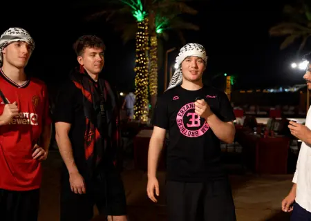At night in what appears to be a desert camp, four young men are gathered. The scene is lit by fairy lights strung around palm trees in the background.