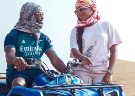 A man and a woman are in a sunny desert setting with a blue quad bike (ATV). The man is sitting on the ATV, wearing a dark teal "Emirates Fly Better" soccer jersey, sunglasses, and a black and white keffiyeh (headscarf). He is looking to his right at the woman, who is standing next to the ATV. She wears a pink and gold patterned headscarf, sunglasses, a light pink t-shirt with a graphic print, and light-colored shorts. She is smiling and pointing towards the front of the ATV.