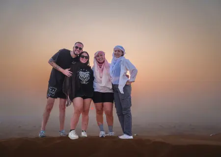 Four people stand close together, posing for a photo on the crest of a sand dune. In the background, a hazy, golden sun sets over the desert.