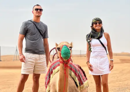 A man in a grey t-shirt and a woman in a white tank top stand on either side of a camel, which is sitting in the desert. The camel wears a colorful red-and-green harness and bridle.
