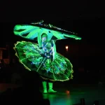 trawey tours A Tanoura dancer spinning during a night performance, wearing a traditional skirt costume illuminated with bright green LED lights, while audience members watch from the dark foreground.