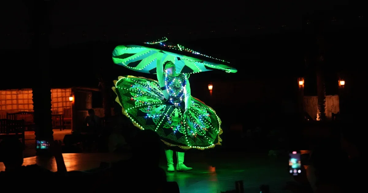 trawey tours A Tanoura dancer spinning during a night performance, wearing a traditional skirt costume illuminated with bright green LED lights, while audience members watch from the dark foreground.