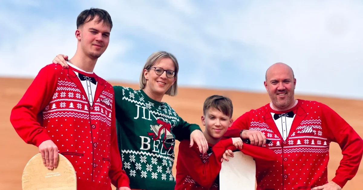 A happy family wearing festive Christmas sweaters and holding a sandboard poses in the Dubai desert dunes during a Trawey Tours holiday safari.