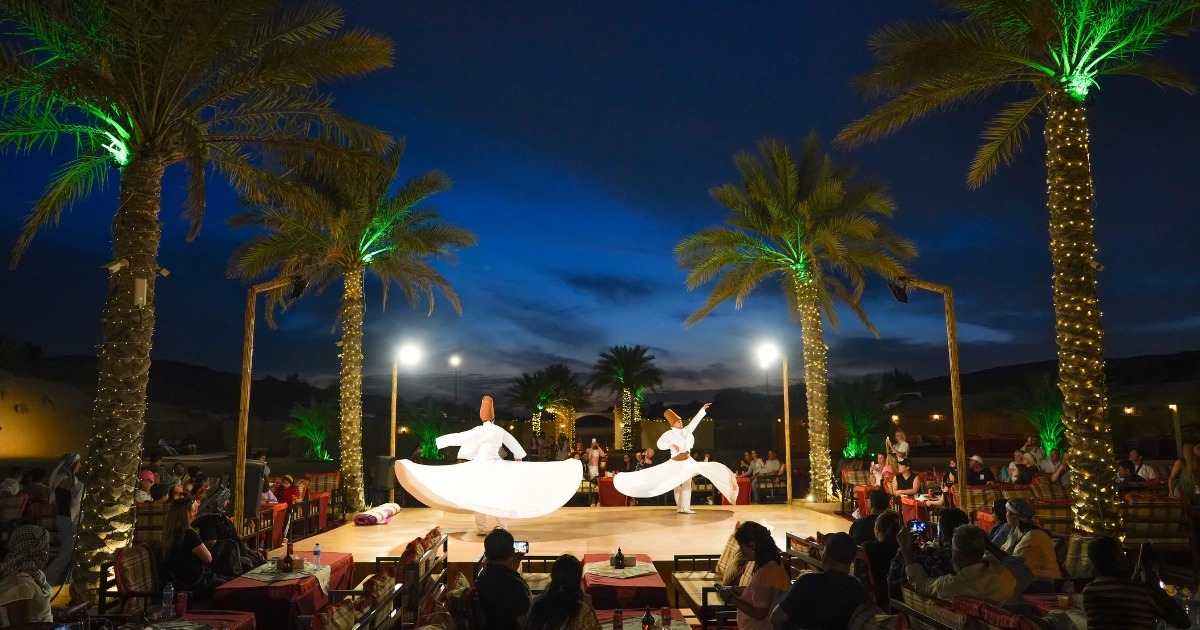 A Tanoura dancer spinning in a vibrant, multi-colored costume on stage at a Trawey desert safari camp in Dubai, viewed by an audience under string lights.