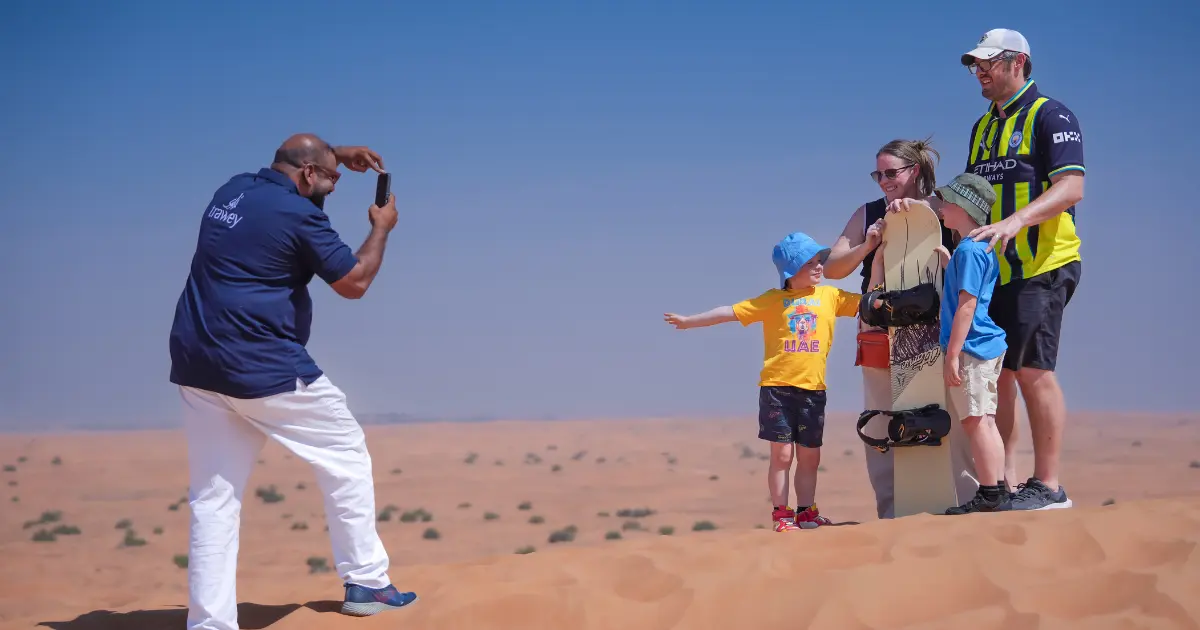 Trawey tour guide taking a photo of a family dressed in light casual clothes holding a sandboard during a Dubai desert safari.