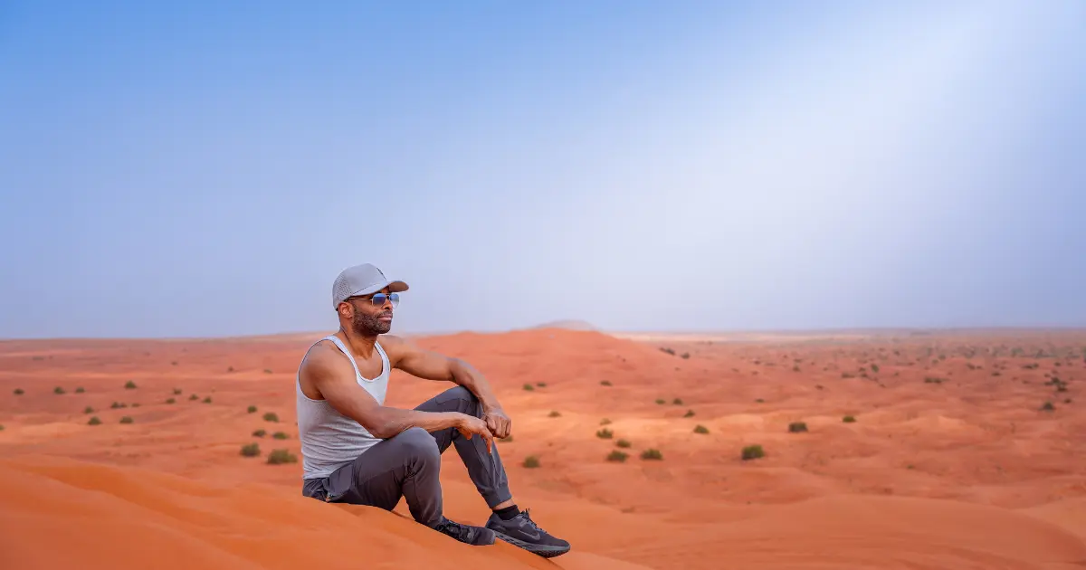 A tourist in a grey tank top sits on a sand dune, enjoying the panoramic view of the Dubai desert landscape during a safari.